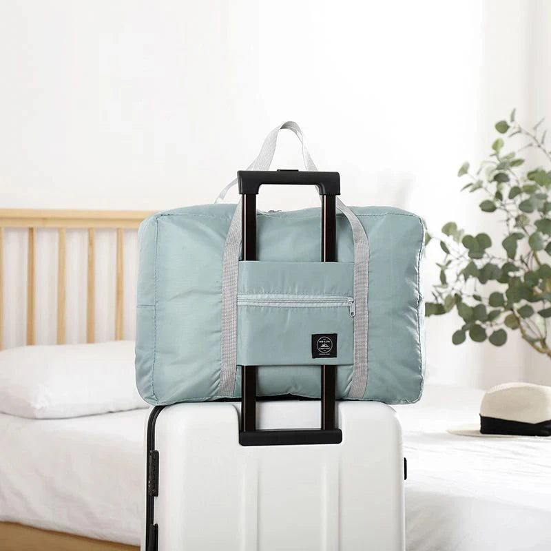 Light blue folding travel bag on a suitcase with a bed and plant in the background