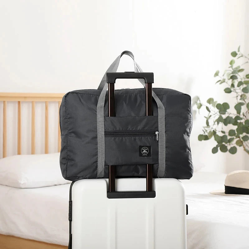 Black folding travel bag with handle on a white suitcase in a bedroom setting
